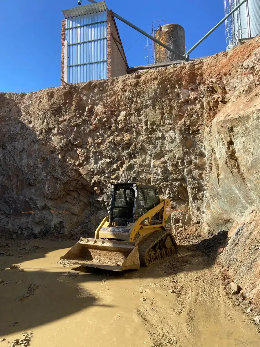 Minicargadora amarilla en un pozo de construcción, trabajando cerca de un edificio con silos, bajo un cielo azul claro.