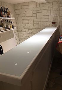 Bar blanc à la surface brillante, adossé à un mur de pierre. Des étagères à bouteilles se trouvent à gauche.