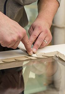 Dans un atelier, des mains positionnent de petites pièces beiges sur une surface vitrée, probablement pour encadrer un objet.