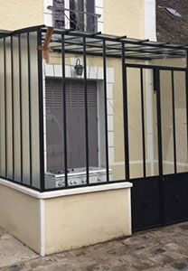 Encadrement en verre et métal noir fixé à un bâtiment couleur crème, avec une porte et une fenêtre.