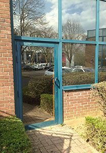 Porte vitrée à cadre bleu, ouverte, encastrée dans un mur de briques. Vue extérieure d'un parking.