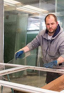 Un homme portant un sweat à capuche et des gants travaille avec du verre et un cadre de fenêtre à l'intérieur.