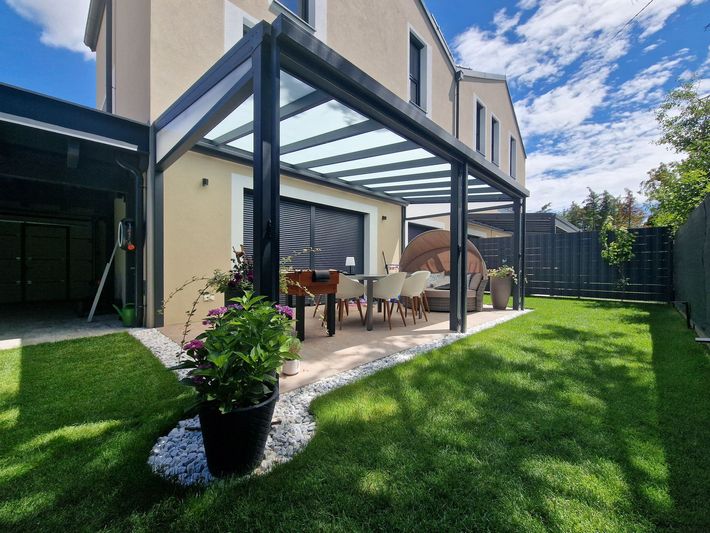 Eine moderne Terrasse mit Glasdach, Tisch und Stühlen, auf einer grünen Wiese, neben einem Haus mit blauem Himmel.