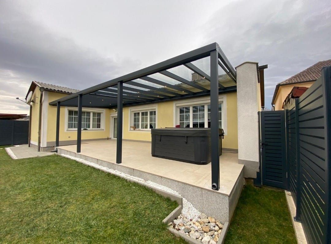 Eine moderne Terrasse mit einer Pergola mit schwarzem Glasdach, einem Whirlpool und einem gelben Haus.