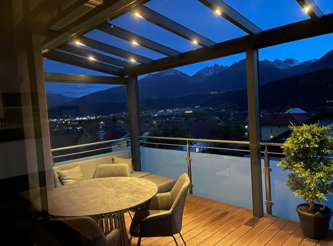 Balkon in der Abenddämmerung mit Blick auf eine Stadt mit Bergkulisse; beleuchteter Tisch und Sitzgelegenheiten.
