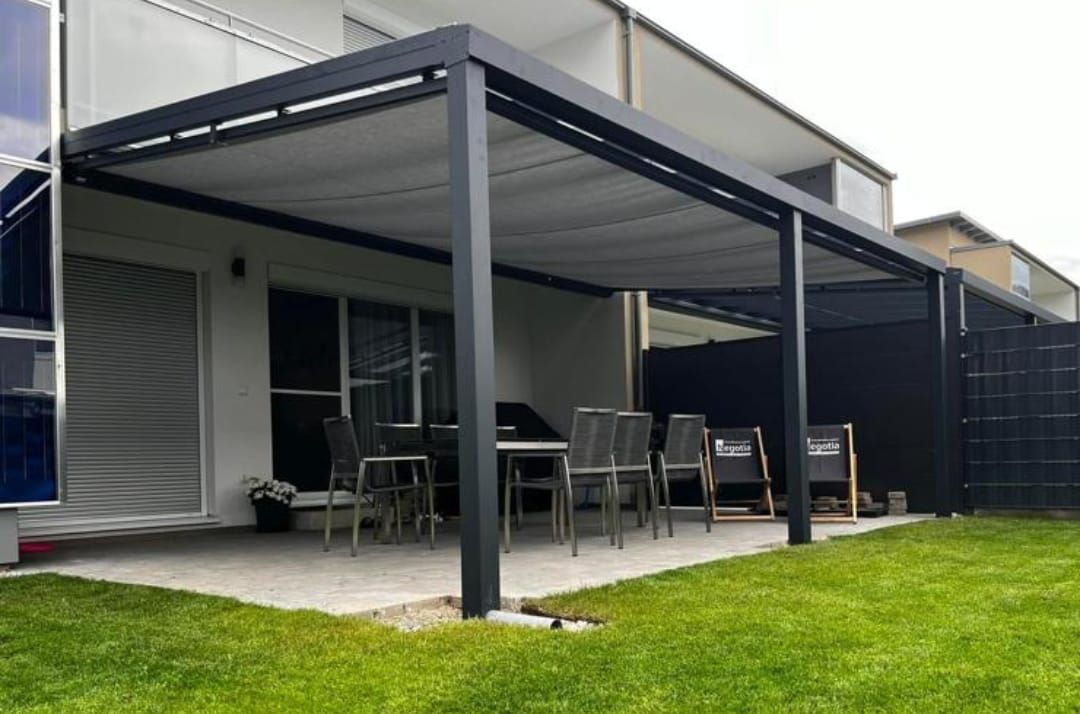 Außenterrasse mit schwarzer Pergola über einem Essbereich, neben einem Haus auf einem grünen Rasen.