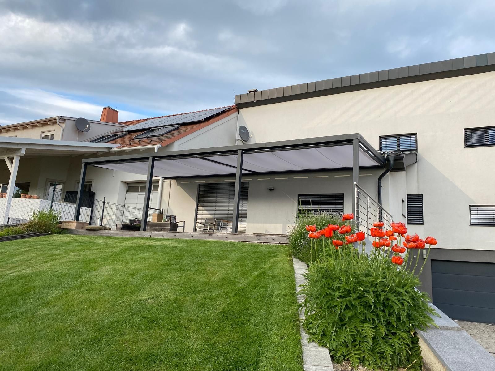 Hinterhof mit grauer Terrassenüberdachung, grünem Rasen, roten Blumen und einem hellen Haus unter einem bewölkten Himmel.