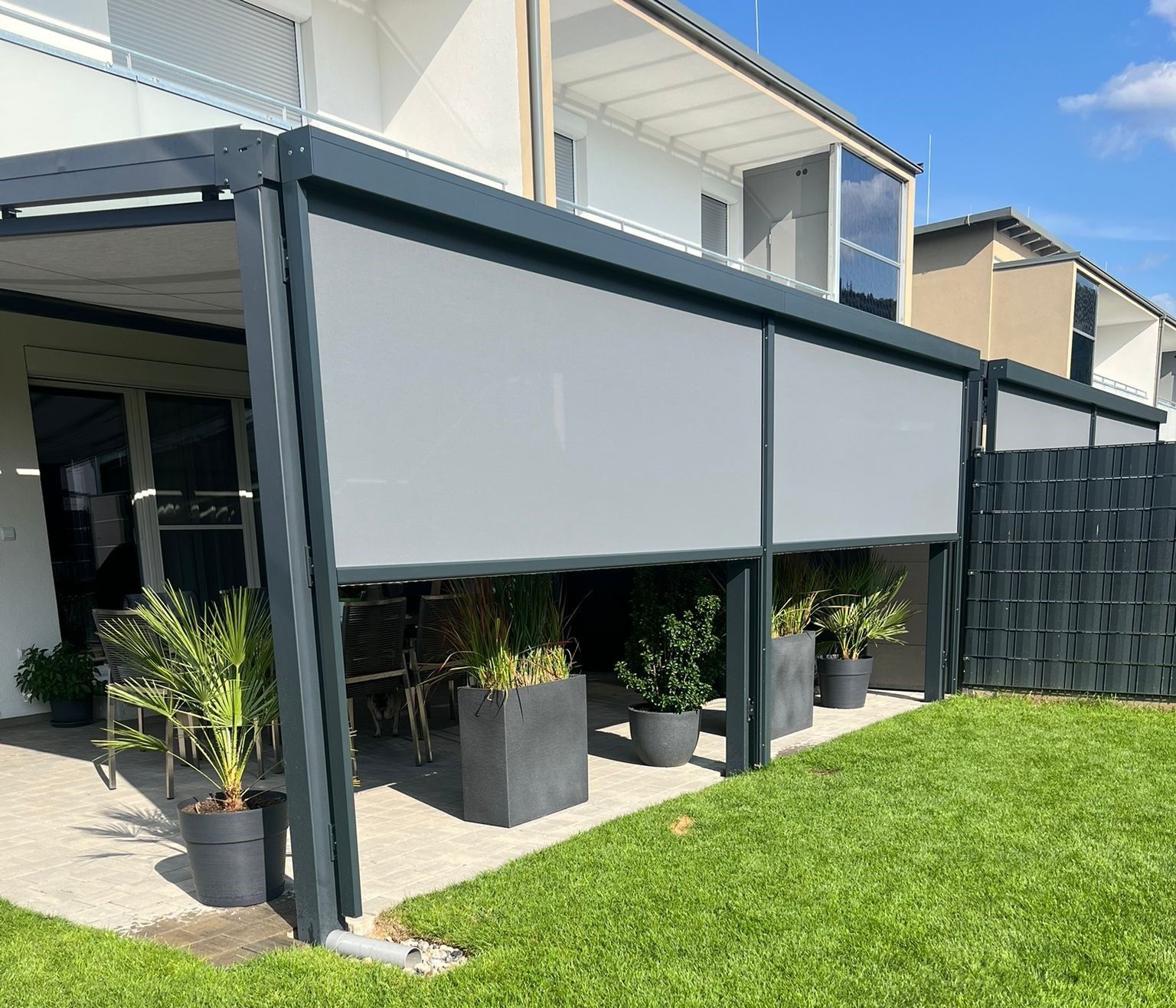 Graue Pergola im Freien mit einziehbaren Sonnenschutzblenden auf einer Terrasse mit Topfpflanzen.