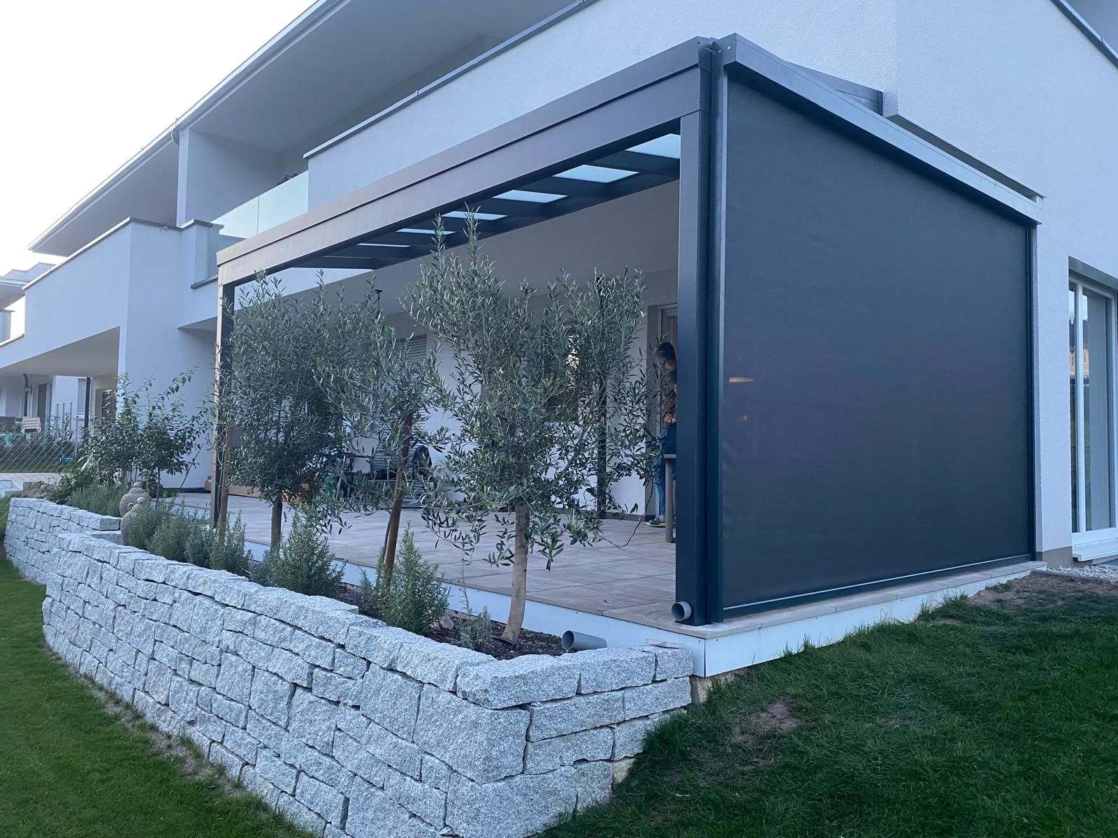 Moderne Terrasse mit ausfahrbarer schwarzer Markise und Seitenmarkise, heller Steinmauer und kleinen Bäumen.