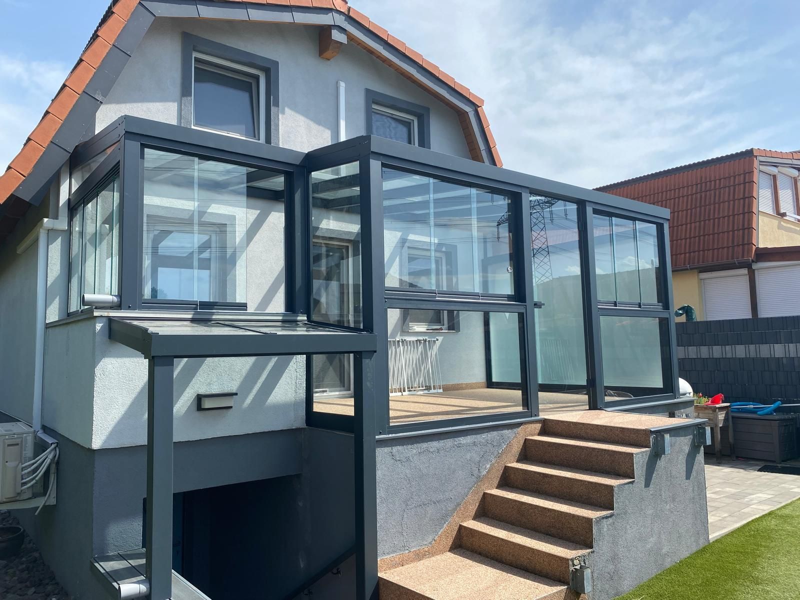 Grauer Wintergartenanbau mit Glaswänden, über eine Treppe an ein hellgraues Haus mit rotem Dach angeschlossen.
