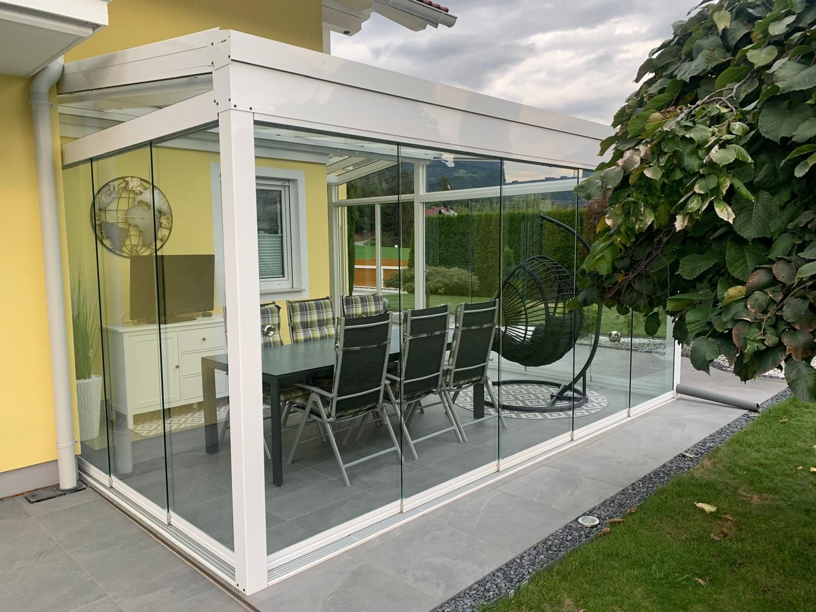 Wintergarten mit weißem Rahmen und Glaswänden, Esstisch und Stühlen, neben einem Haus und Garten.