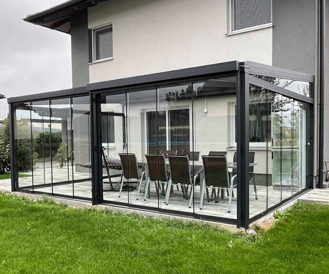 Verglaste Terrasse mit Esstisch und Stühlen, an ein hellgraues Haus angeschlossen. Schwarzer Rahmen, grünes Gras.
