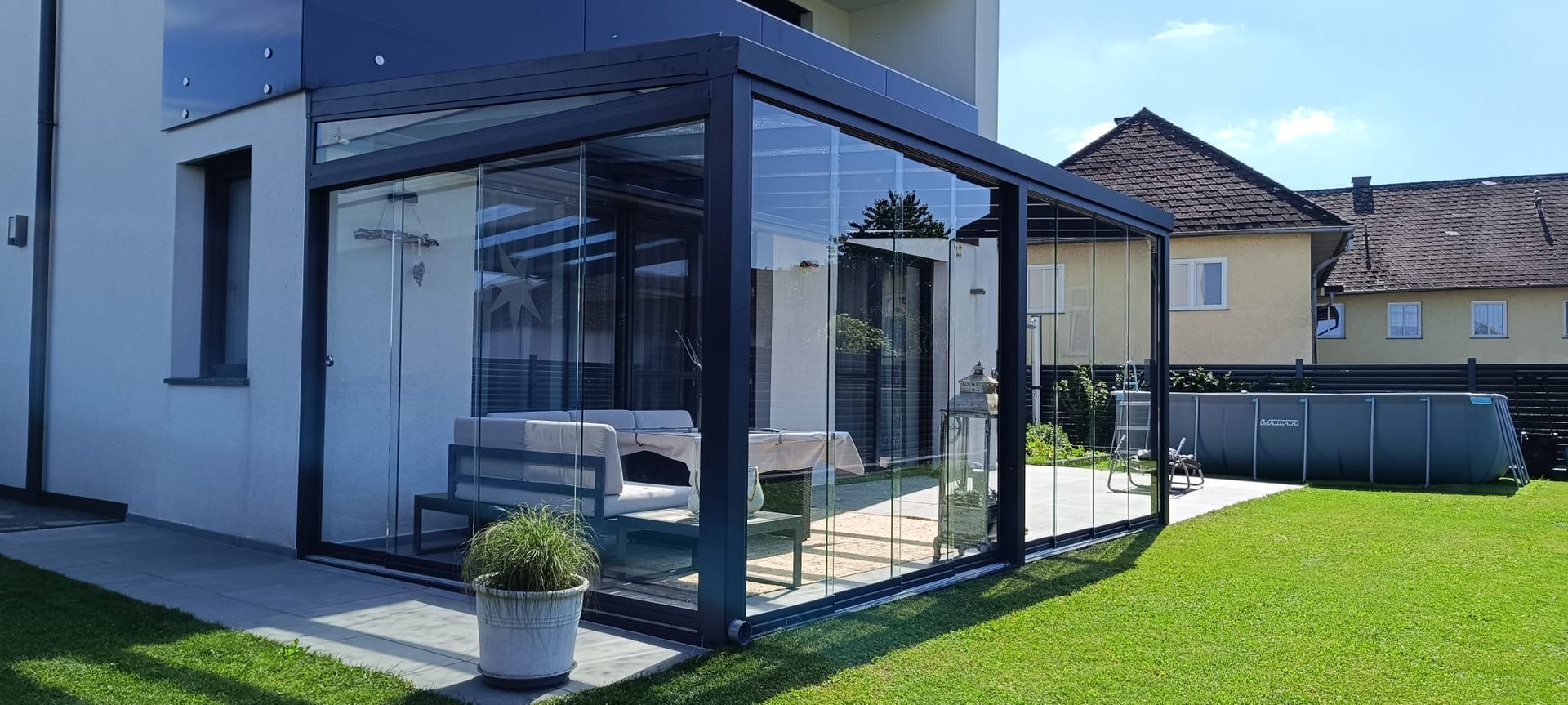 Eine moderne, verglaste Terrasse mit Sitzgelegenheiten, neben einem weißen Haus, auf einer Rasenfläche.
