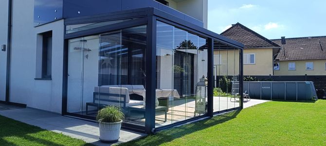 Eine moderne, verglaste Terrasse mit Sitzgelegenheiten, neben einem weißen Haus, auf einer Rasenfläche.