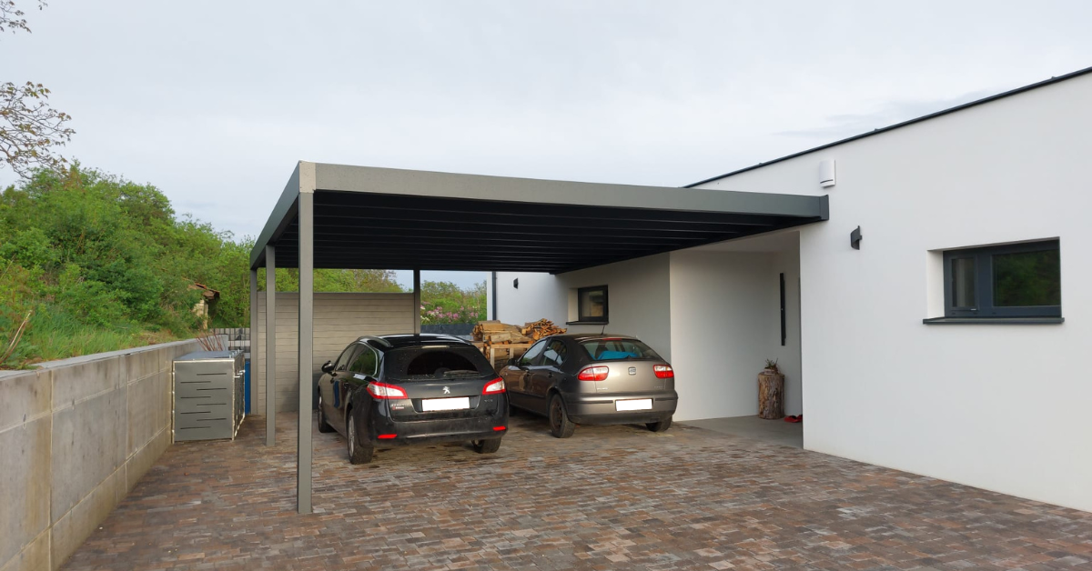 Schwarzer Carport auf gepflasterter Fläche, mit Flachdach. Graue Steinterrasse. Grüne Sträucher.