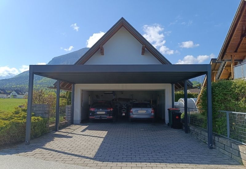 Carport über der Auffahrt, die zu einer Garage mit zwei darin geparkten Autos führt. Berge im Hintergrund.