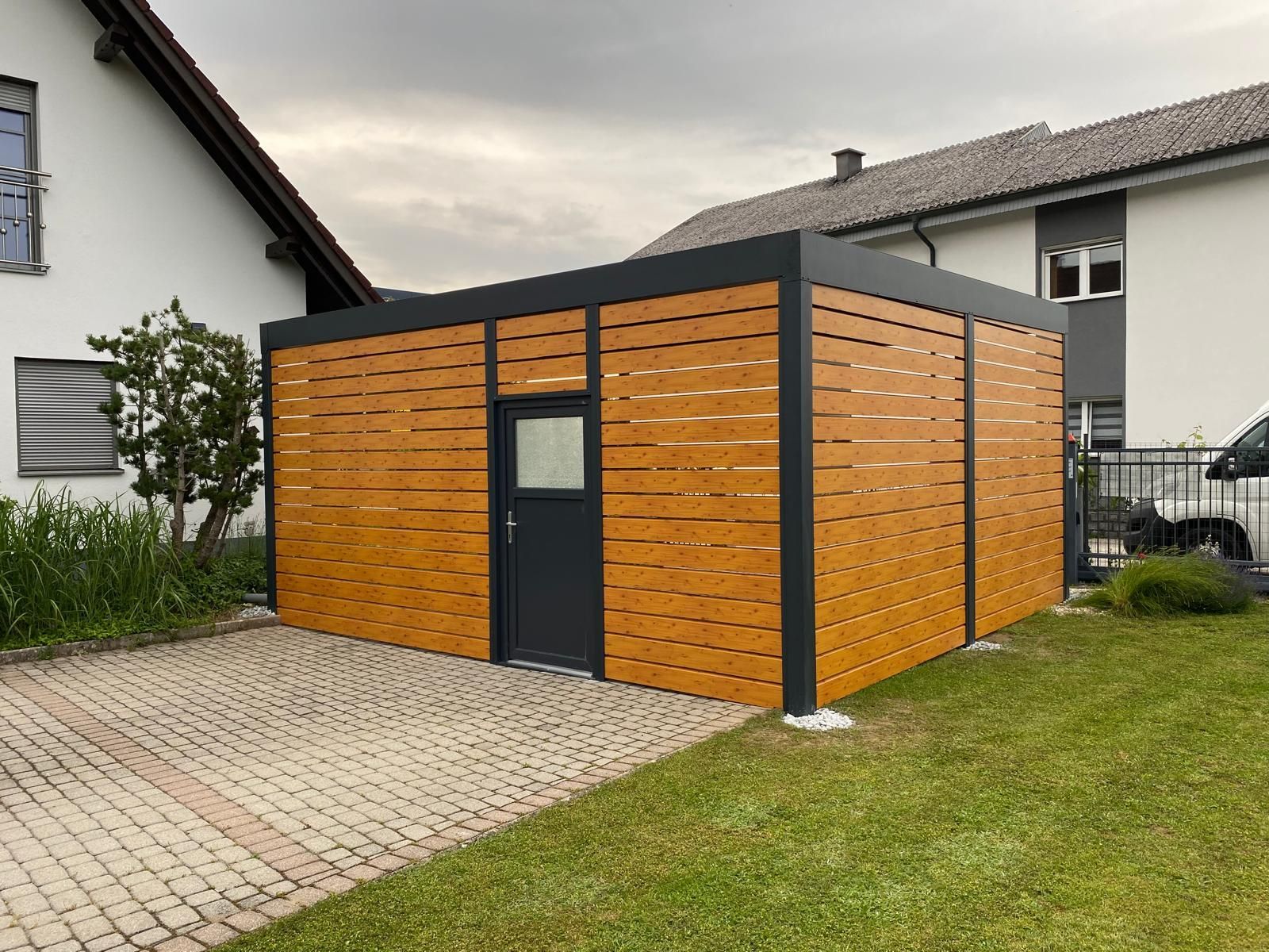 Moderner Carport mit Holzlattenwänden, der ein auf einer gepflasterten Auffahrt neben einem Haus geparktes Auto überdeckt.