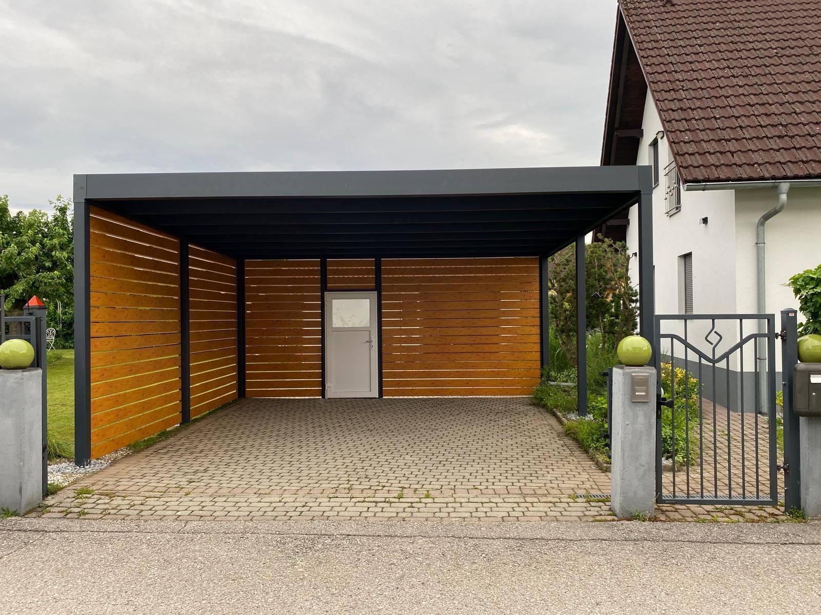 Carport mit Holzlattenwänden, dunklem Dach, heller Tür. Auffahrt mit Kopfsteinpflaster, im Hintergrund Haus.