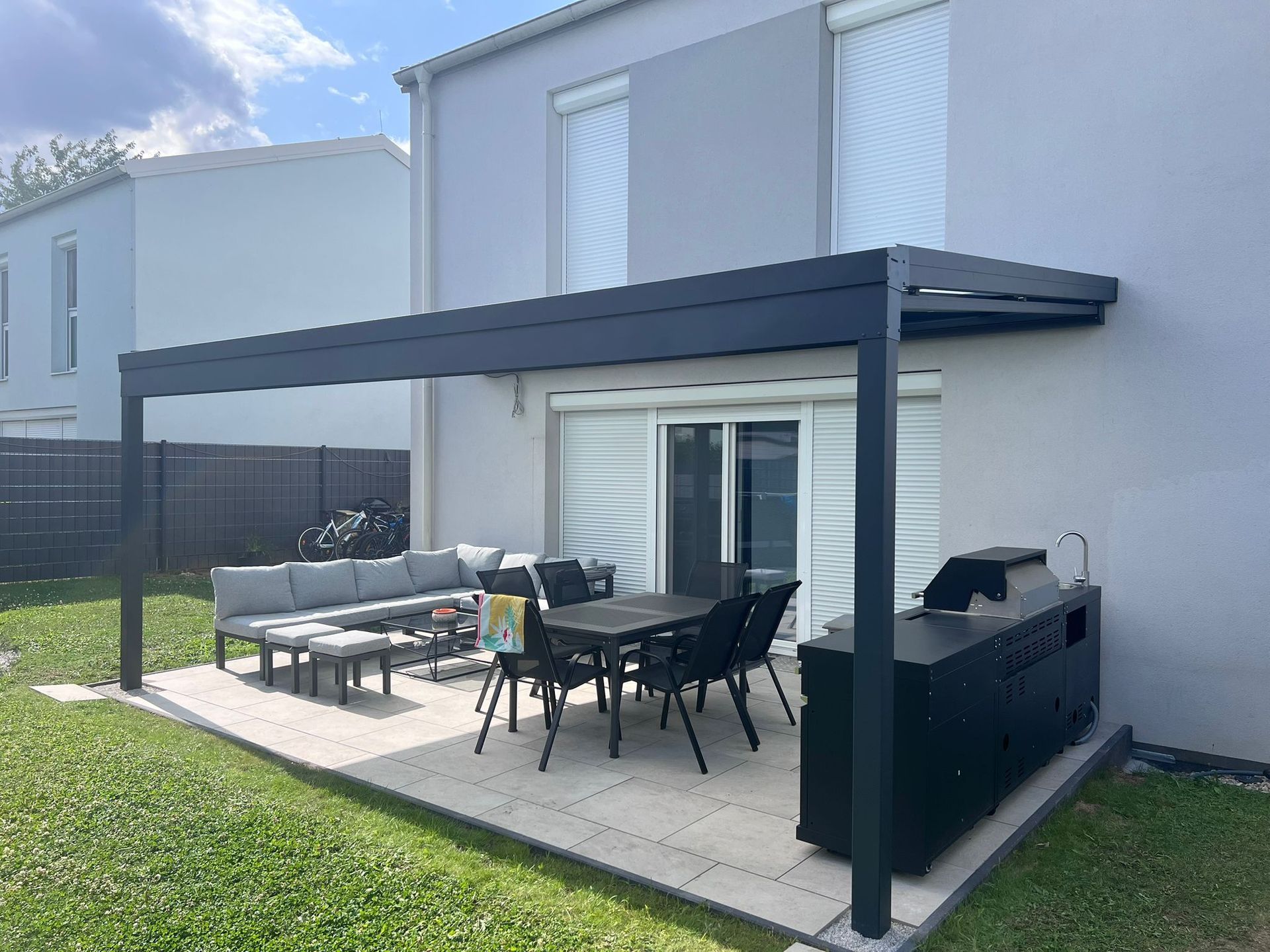 Außenterrasse mit Pergola, Sitzgelegenheiten und Grill neben einem hellgrauen Haus an einem sonnigen Tag.