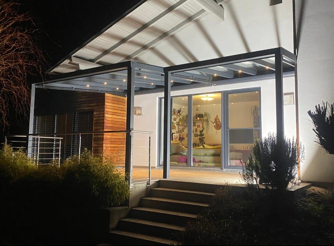 Nachtansicht einer Terrasse mit Pergola, Treppe und beleuchtetem Innenraum.