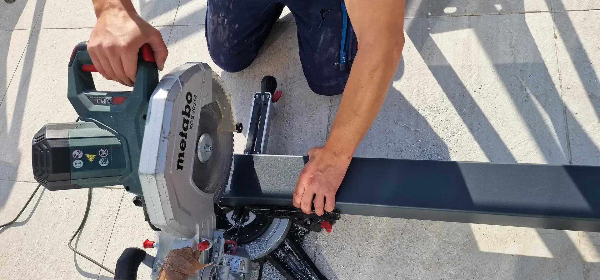 Mann installiert Verbunddeck mit braunen Brettern, Befestigungselementen und einer Bohrmaschine in einer sonnigen Umgebung im Freien.