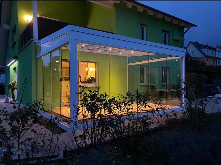 Glass-walled sunroom attached to a green house, lit up at dusk.