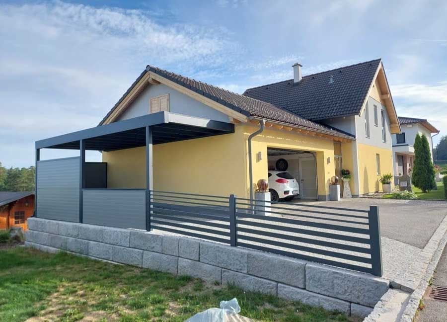 Modern home with attached garage, carport, and gray fence; yellow exterior, blue sky.