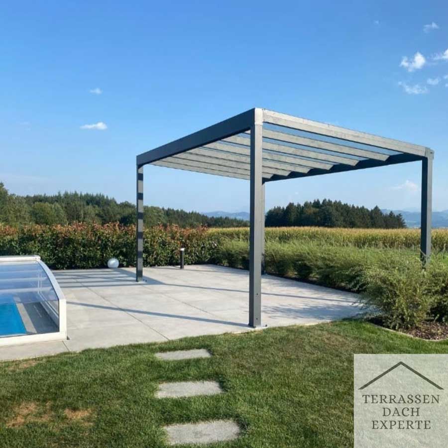 Modern grey pergola on a concrete patio overlooking fields and trees on a sunny day.
