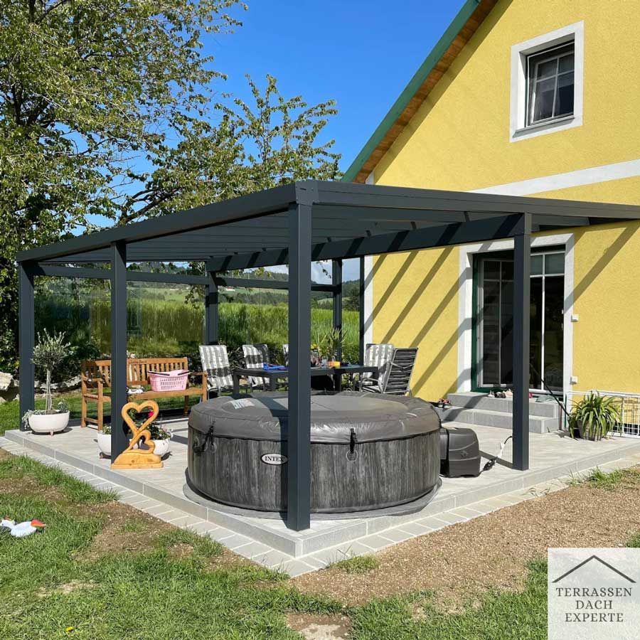 Outdoor patio with dark gray pergola, hot tub, and seating next to a yellow house in a yard.