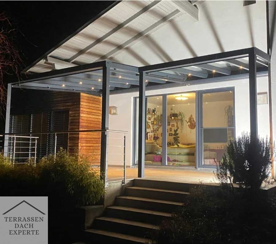 Terrace with lights at night. Modern gray pergola, glass doors, steps, and a wooden facade.