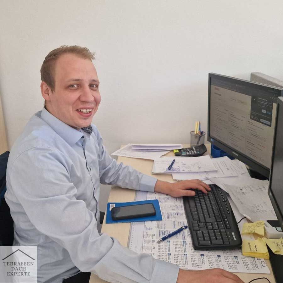 Man smiling at a desk, working on a computer and papers in an office.