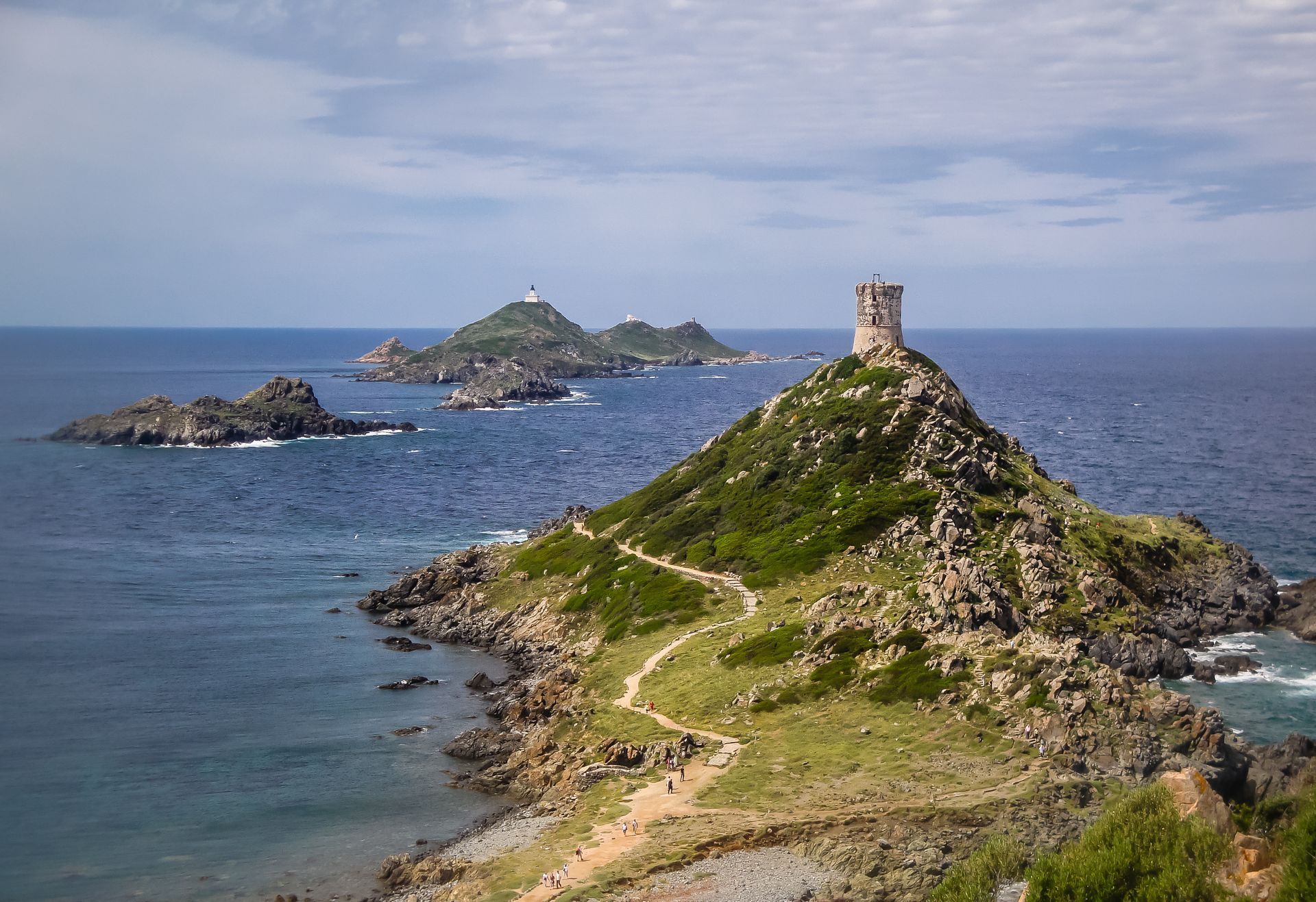 Péninsule côtière rocheuse avec une tour en pierre, des collines verdoyantes et une mer bleue sous un ciel nuageux.