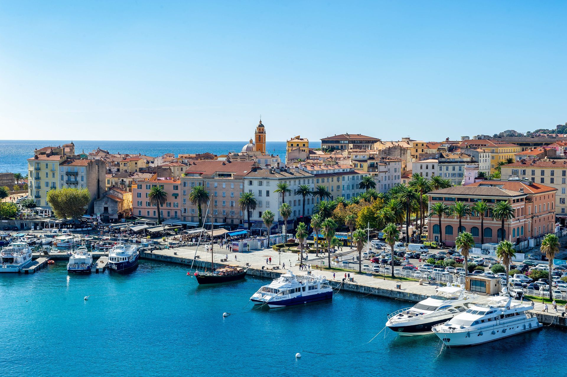 Port ensoleillé avec des bateaux blancs et des bâtiments en bord de mer le long d'une baie bleue.