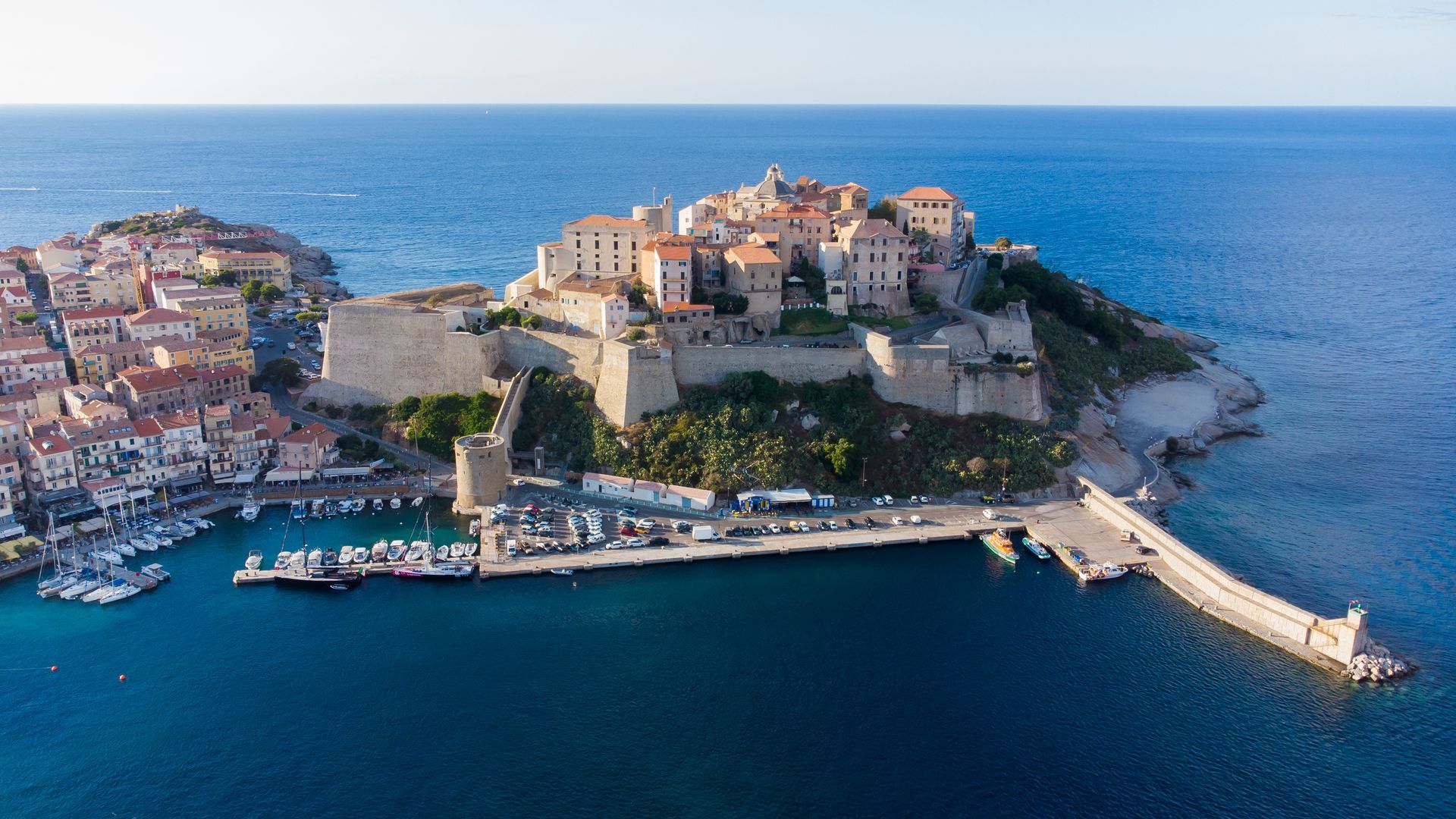Vue aérienne d'un port côtier avec une ville historique perchée sur une colline et entourée par la mer bleue.