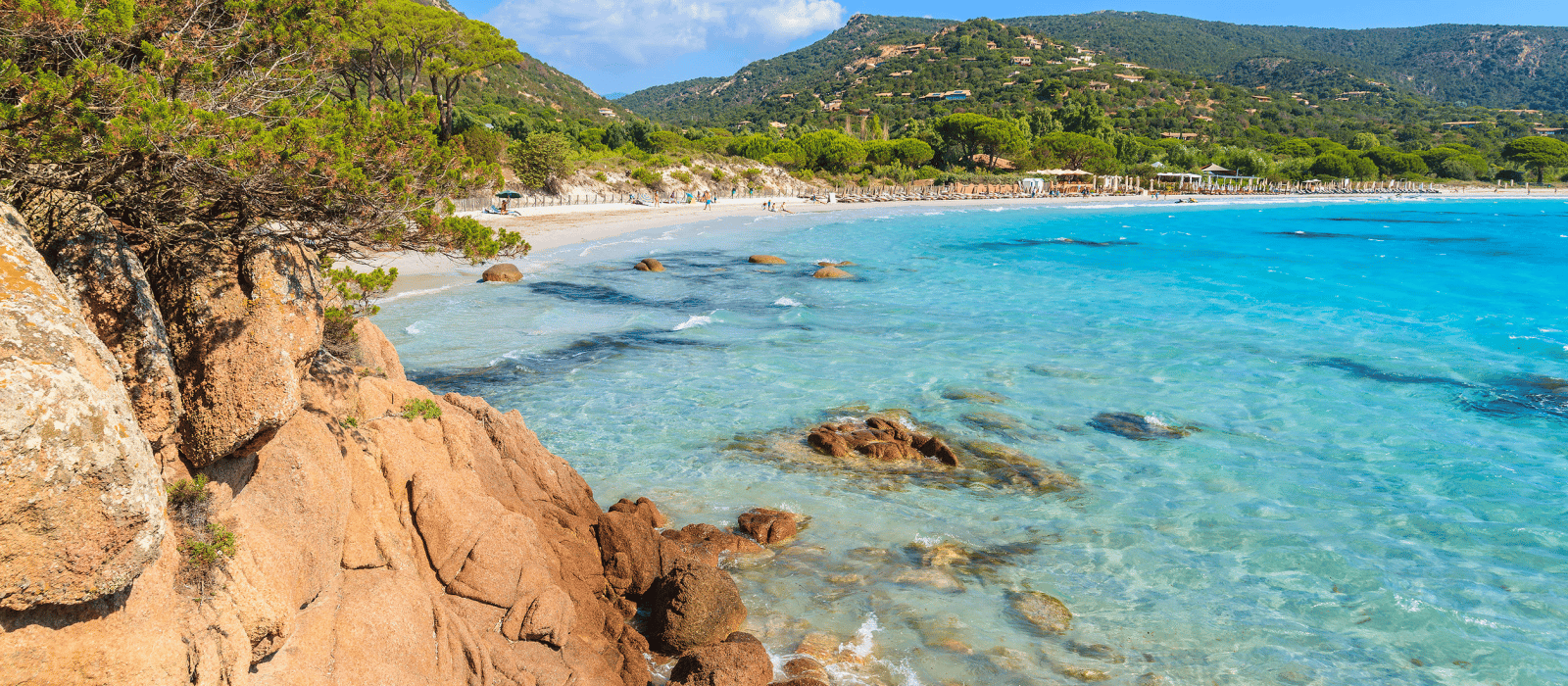 Plage tropicale rocheuse aux eaux turquoise limpides et aux collines verdoyantes sous un ciel ensoleillé.