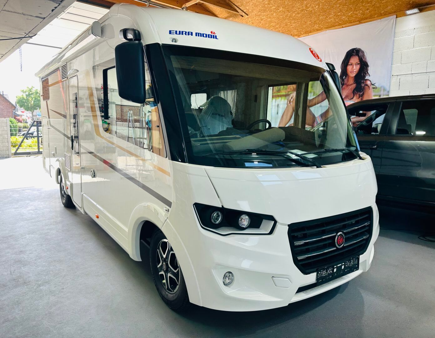 Ein weißes Wohnmobil steht in einem Ausstellungsraum neben einem schwarzen Lieferwagen.