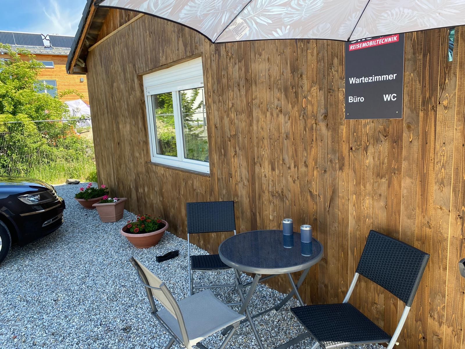 Vor einem Holzgebäude mit einem Tisch und Stühlen unter einem Sonnenschirm ist ein Auto geparkt.