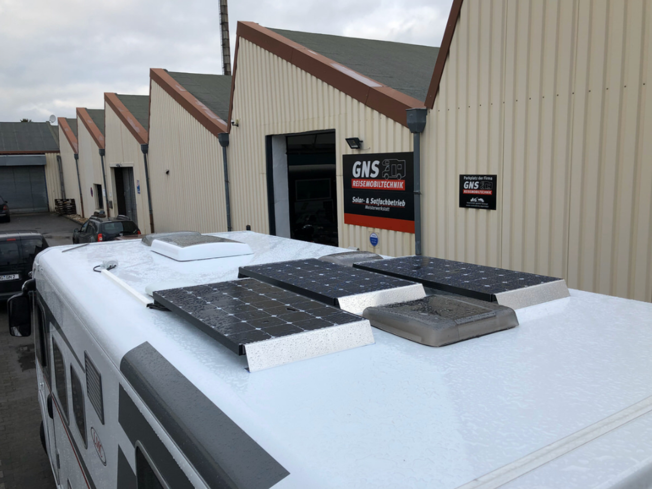 Ein weißer Bus mit Solarzellen auf dem Dach steht vor einem Gebäude.