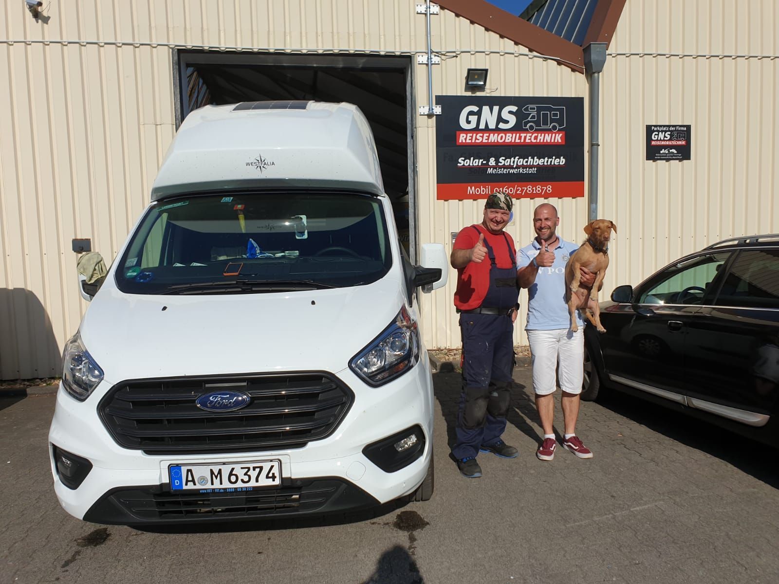 Zwei Männer und ein Hund stehen vor einem weißen Lieferwagen.