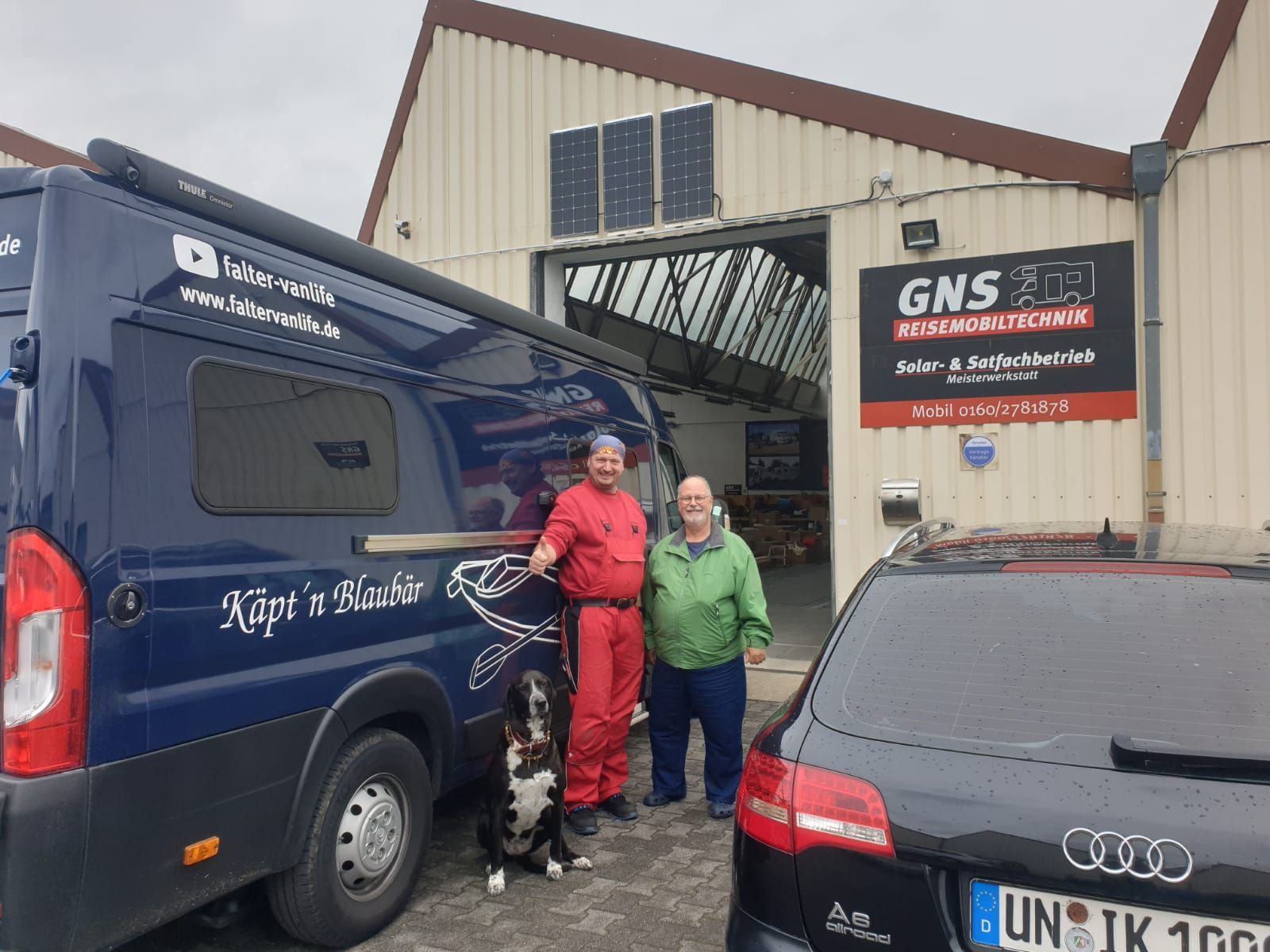 Zwei Männer und ein Hund stehen vor einem blauen Lieferwagen.