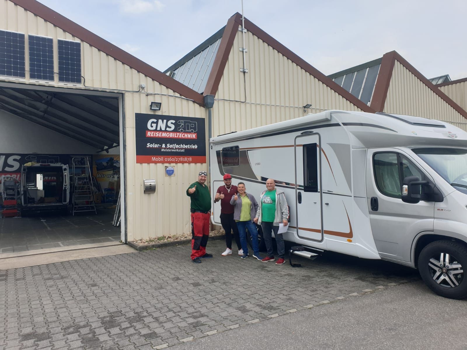 Eine Gruppe von Leuten steht vor einem Wohnmobil, das vor einem Gebäude geparkt ist.