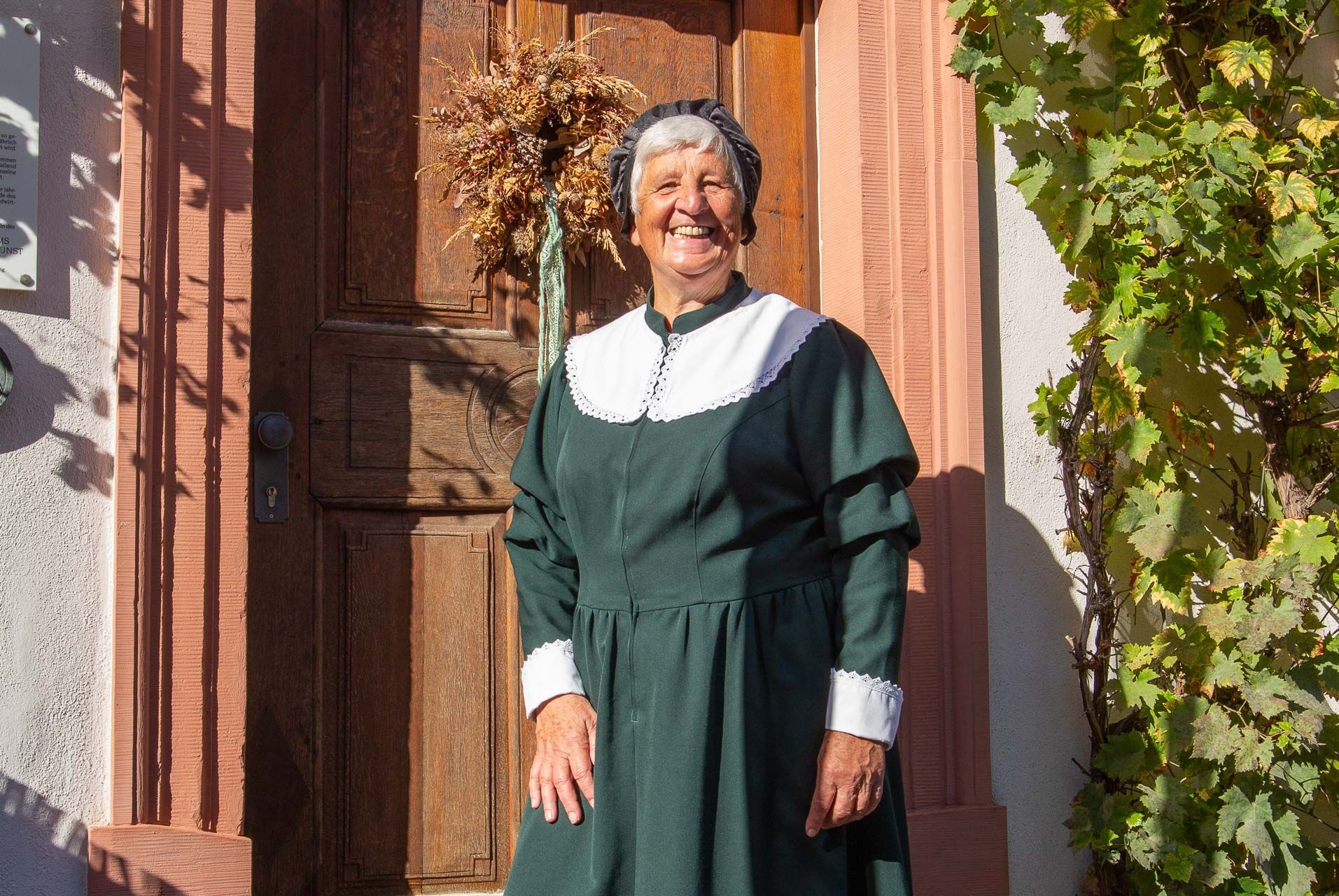 Eine lächelnde Frau in einem dunkelgrünen Kleid mit einem weißen Kragen und einer Haube auf dem Kopf steht vor einer Holztür.