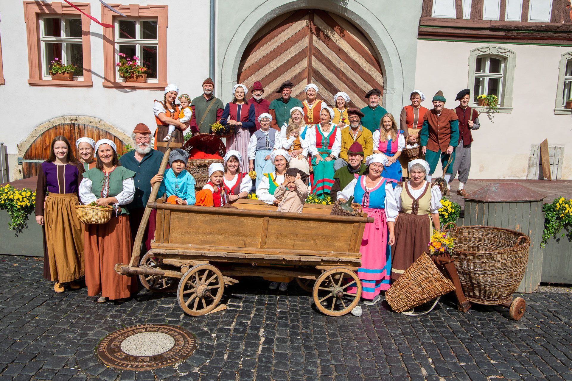 Eine große Gruppe in historischen Kostümen posiert vor einem steinernen Torbogen, einige Leute sitzen in einem Holzwagen.