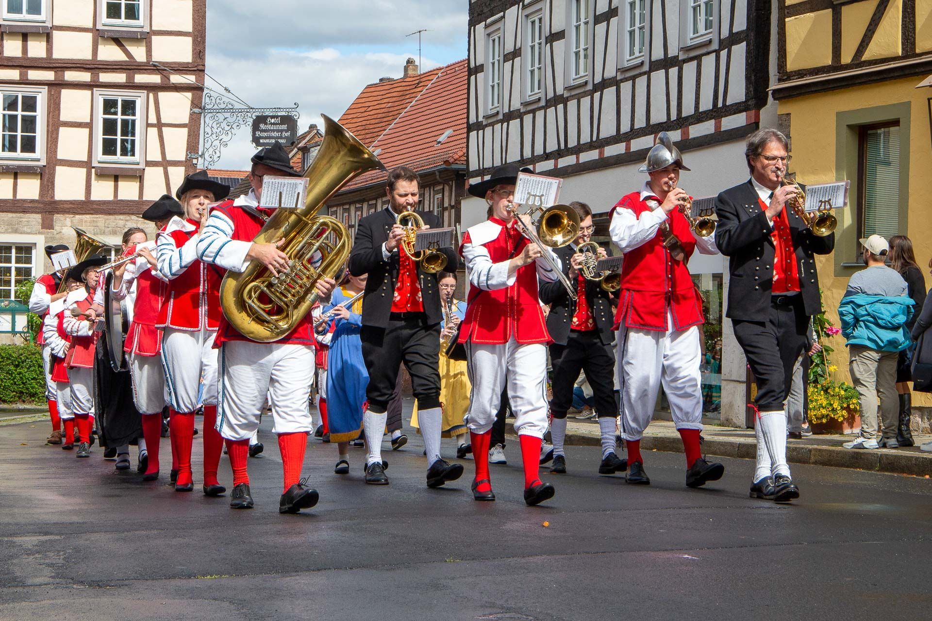 Eine Blaskapelle in rot-weißen Uniformen spielt Blechblasinstrumente auf einer Straße, die von Fachwerkhäusern gesäumt ist.