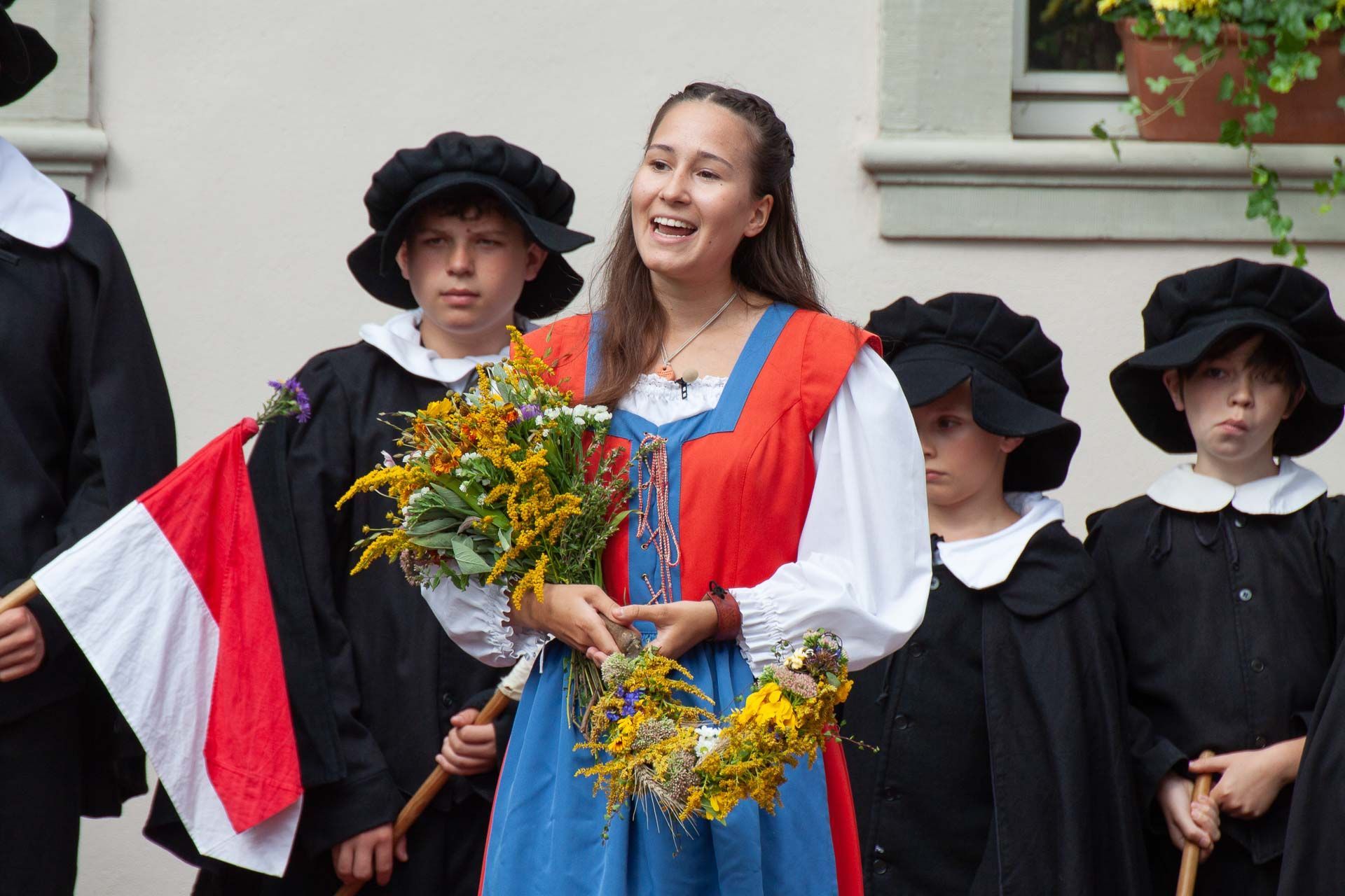 Szene aus dem Heimatspiel mit einer jungen Frau in buntem Kleid, dahinter eine Gruppe Jungen in schwarten Umhängen und Mützen