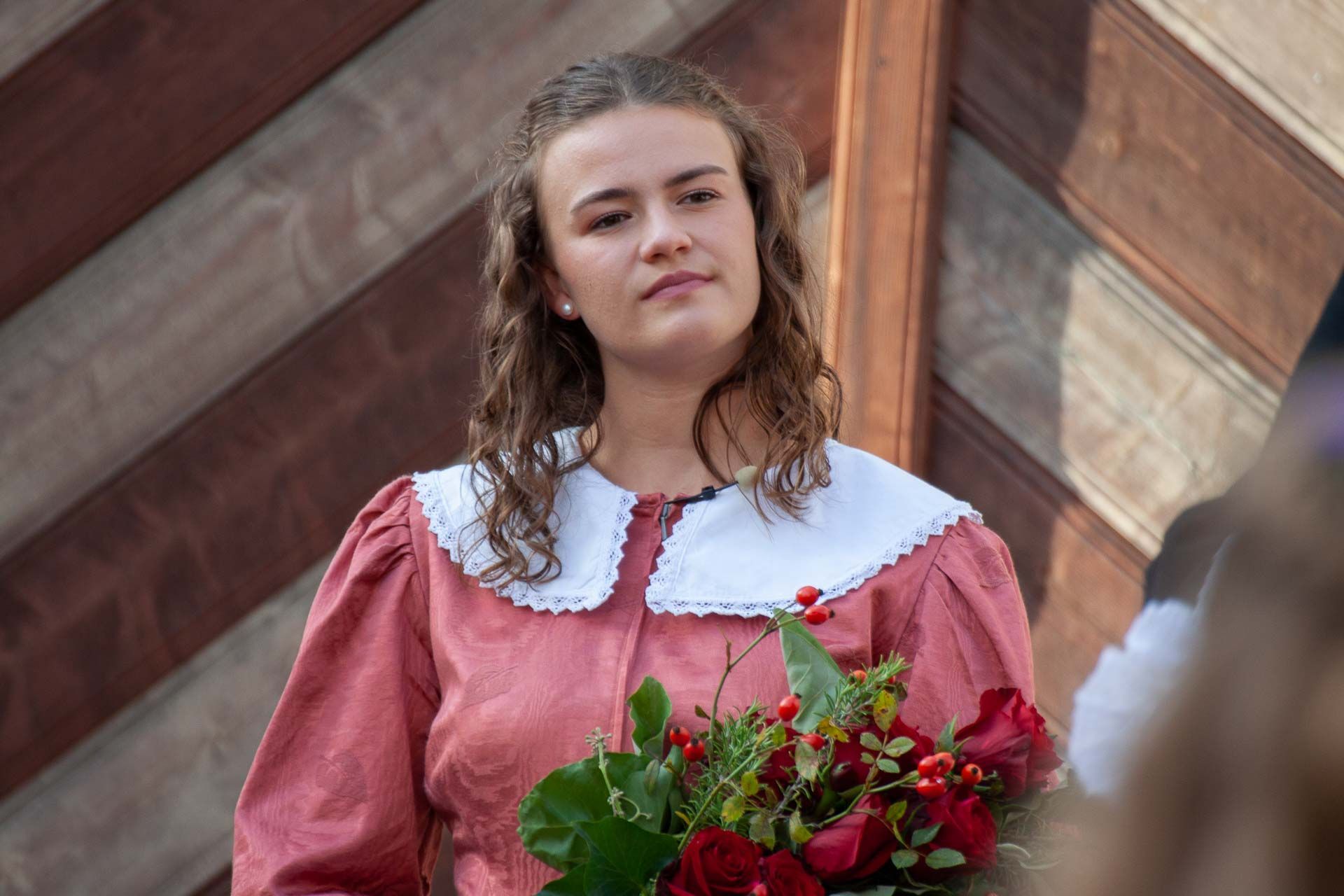 Eine junge Frau mit rosa Kleid und weißem Kragen steht vor einem Holztor