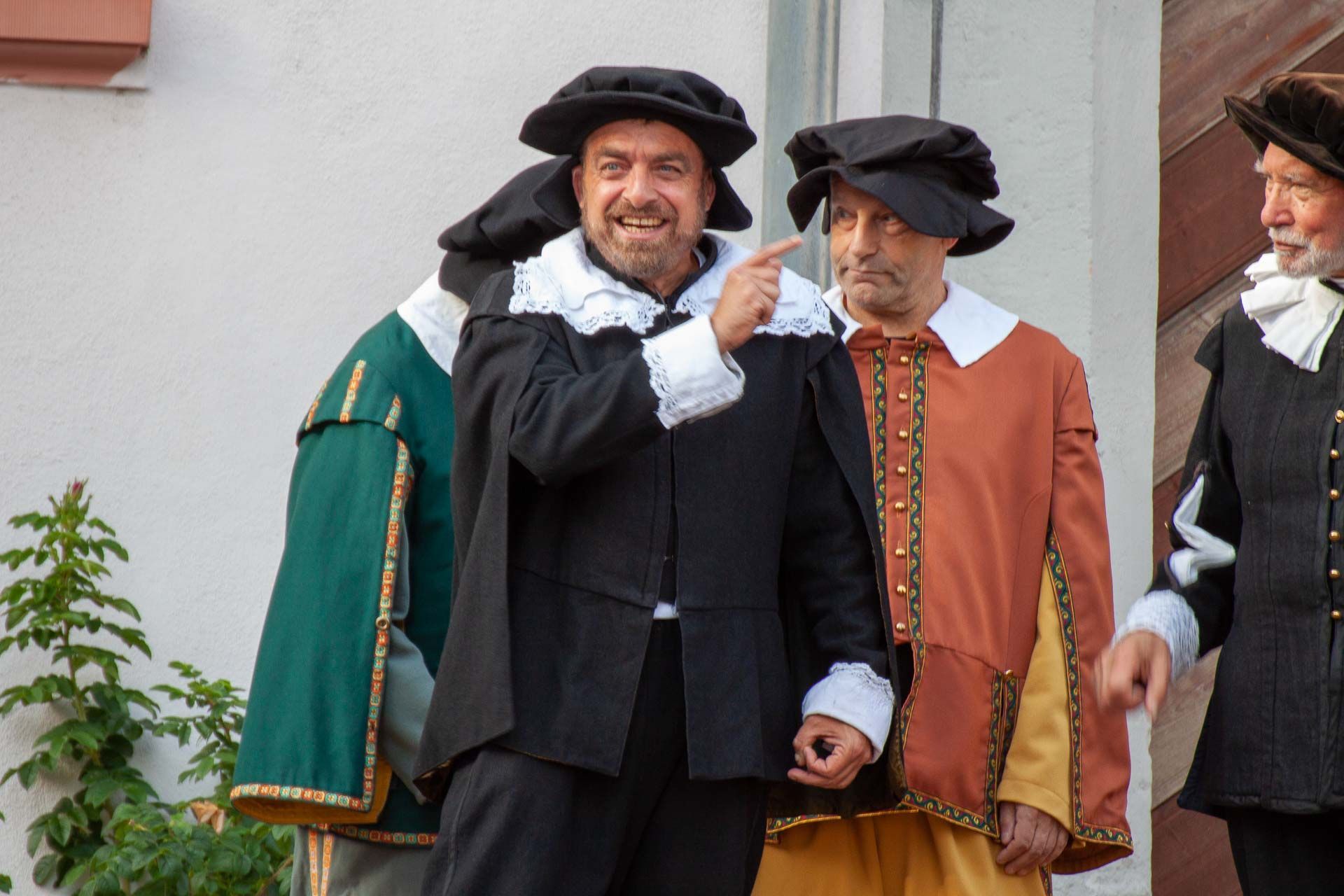 Ein Mann in schwarzem Kostüm mit weißem Kragen und Hut gestikuliert, dahinter weitere Männer in historischen Gewändern