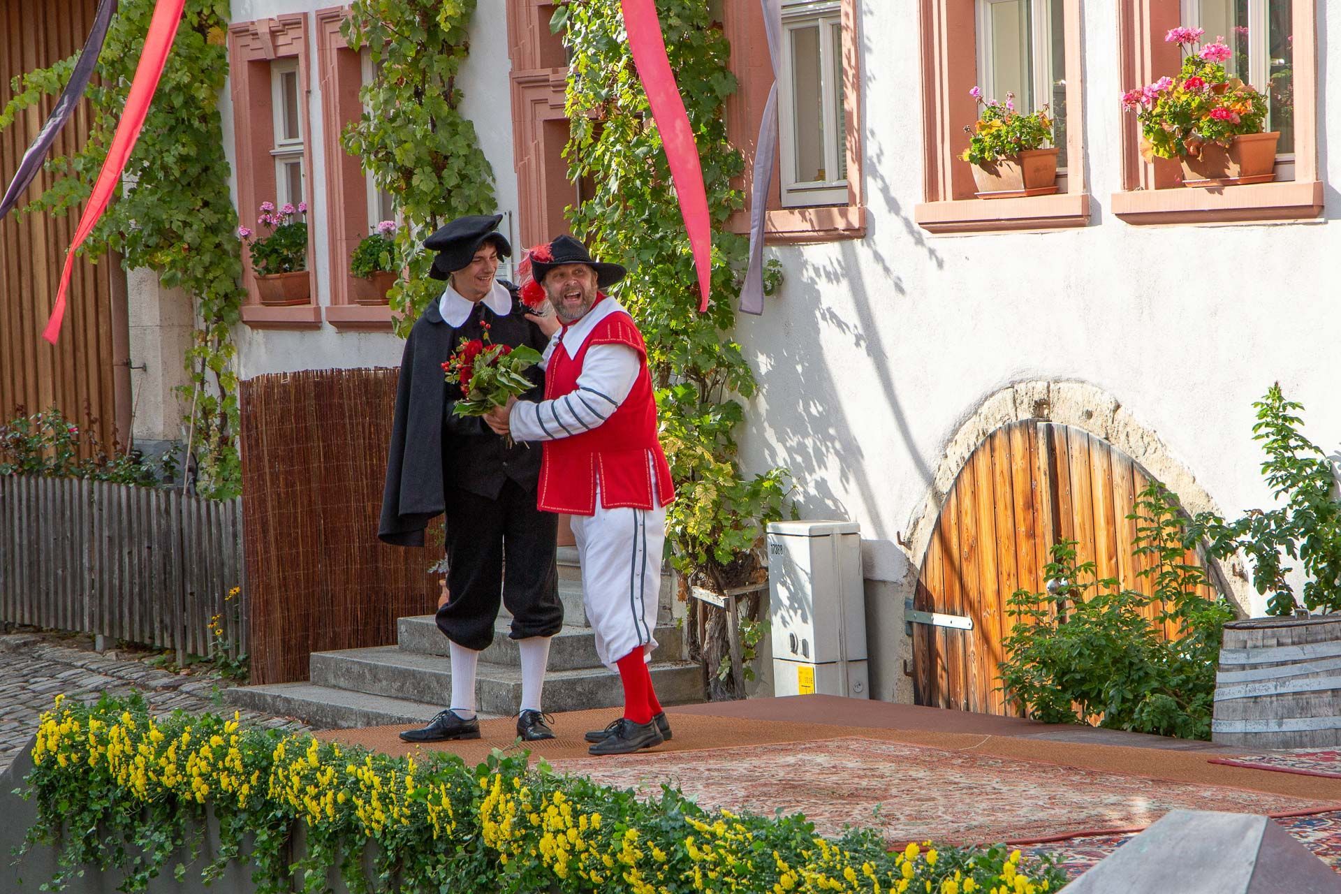 Szene aus dem Heimatspiel mit zwei Männern im Gespräch, einer hält einen Strauß Blumen in der Hand