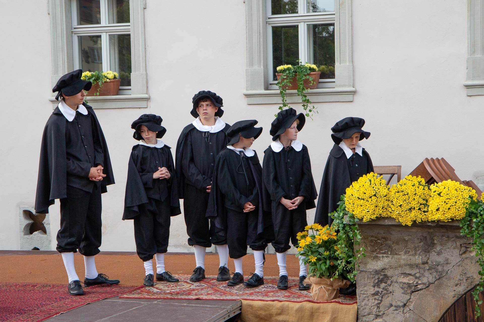 Szene aus dem Heimatspiel mit Jungen in schwarzen Kostümen mit Umhang, weißem Kragen und Mützen