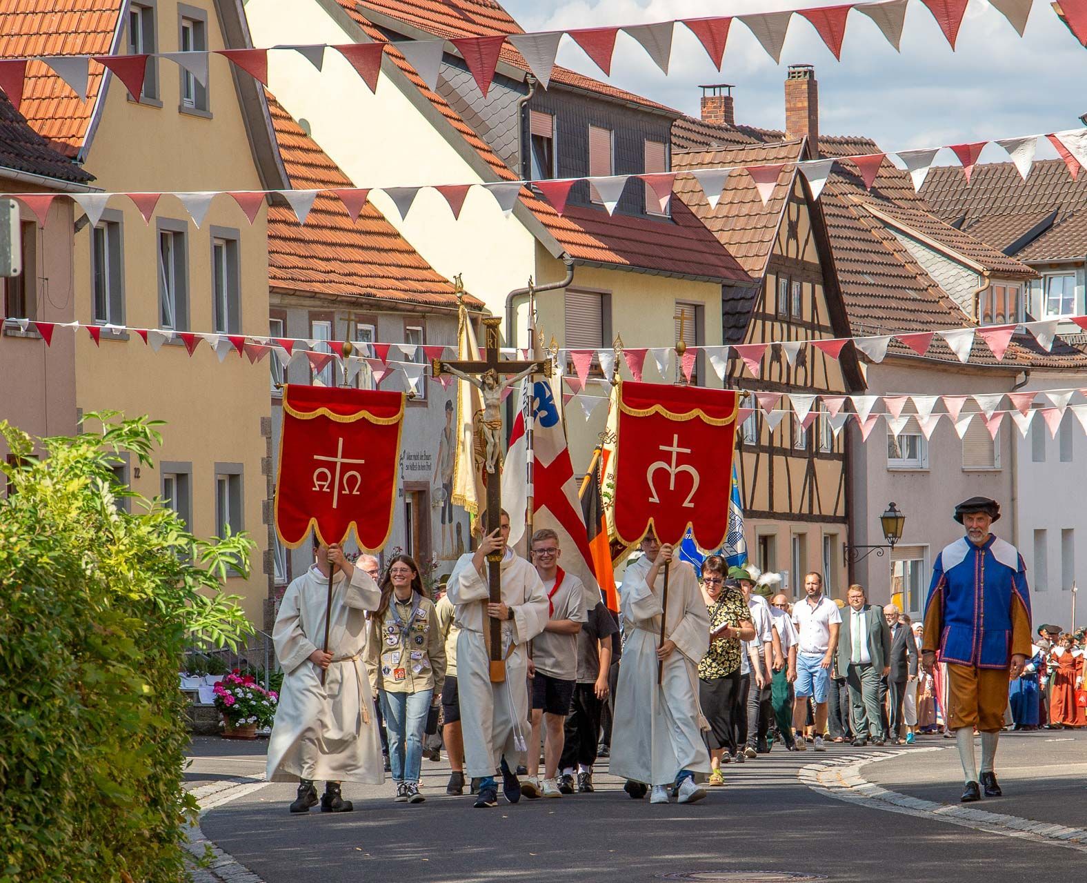 Eine religiöse Prozession zieht mit Bannern und einem goldenen Kruzifix durch eine geschmückte Straße.
